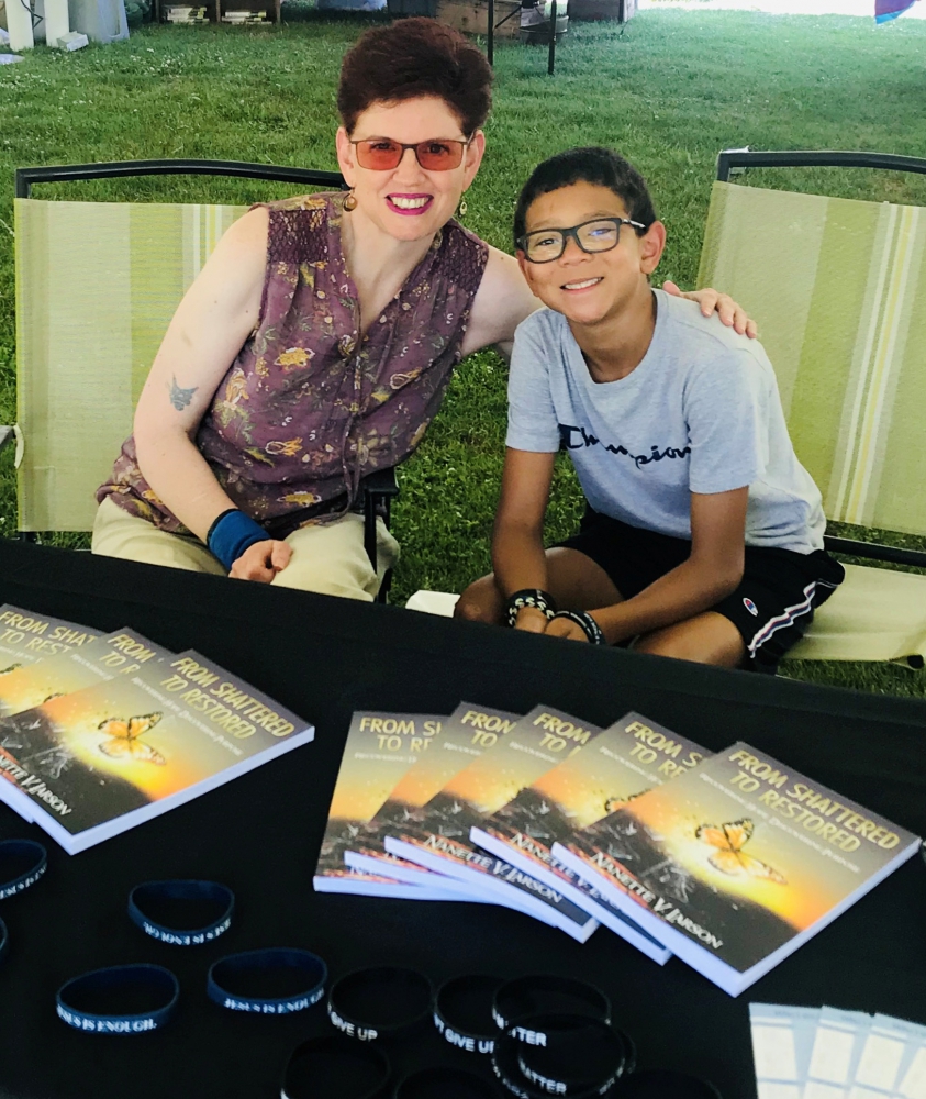 Nannette V. Larson sitting with a kid behind a table with copies of the From Shattered to Restored book